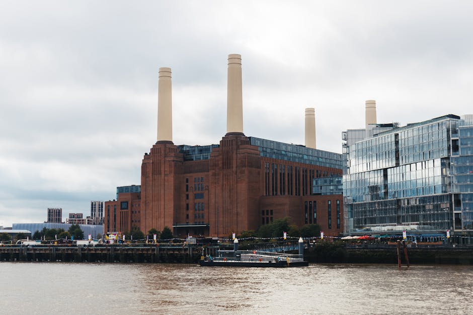 A historic red brick building with four tall, cream-colored smokestacks situated along the riverbank, framed by modern glass-fronted office buildings on the right. The scene is captured under a cloudy