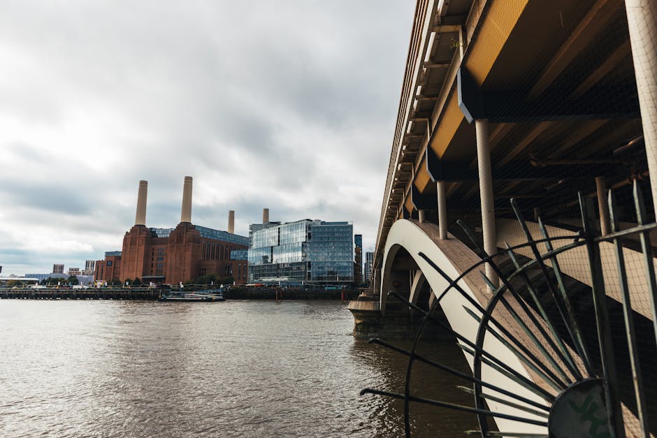 A view of the River Thames in Battersea featuring a modern building with glass facades and four tall chimney stacks of a historic power station in the background, under a cloudy sky. In the foreground
