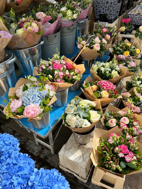 A vibrant display of freshly arranged floral bouquets and potted flower arrangements at a market stall. The bouquets feature a mix of roses, hydrangeas, and lilies in shades of pink, white, and blue, 