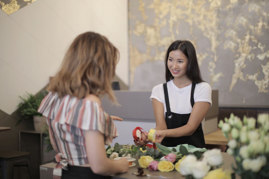 A young female florist with long dark hair, wearing a white t-shirt and black apron, hands a small floral gift with red wrapping and yellow ribbon to a female customer with shoulder-length wavy blonde