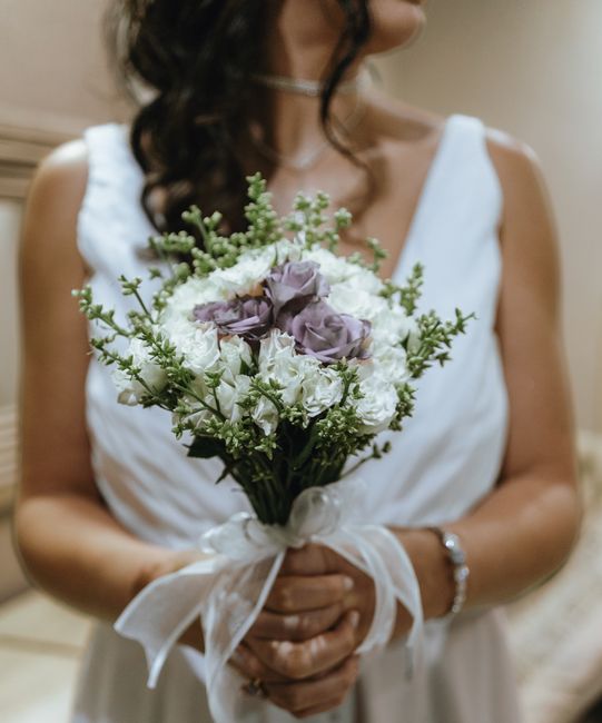 Close-up of a bride holding a floral bouquet, featuring white and light purple roses, complemented by sprigs of green foliage with small white flowers. The bouquet is wrapped with a white satin ribbon