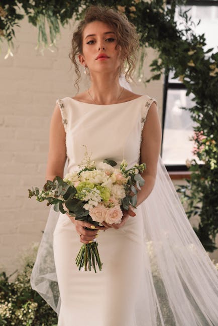 A young woman with curly, shoulder-length hair holds a floral bouquet in a bright indoor setting with natural light streaming through large windows. The bouquet features soft pastel-colored flowers, i